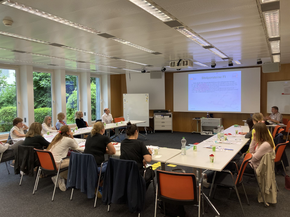 Teilnehmende eines Seminars sitzen in einem rechteckigen Tisch-Arrangement in einem hellen Raum mit grossen Fenstern. Eine Präsentation mit dem Titel «Stolpersteine FI» wird an die Wand projiziert. Die Atmosphäre wirkt aufmerksam und konzentriert.