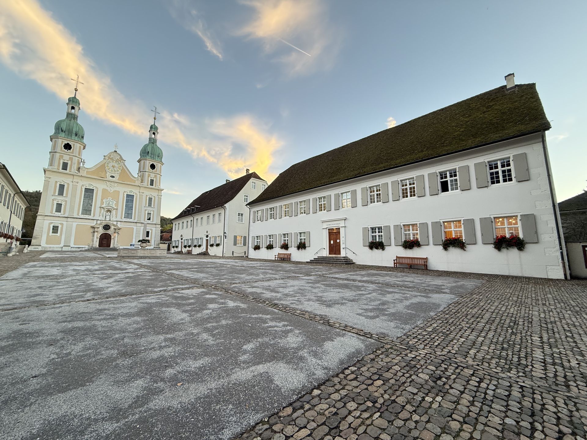 Blick über den Domplatz in Arlesheim bei Sonnenuntergang. Links steht die Domkirche, rechts das historische Gemeindehaus mit seinen hellen Fassaden und Fensterläden.