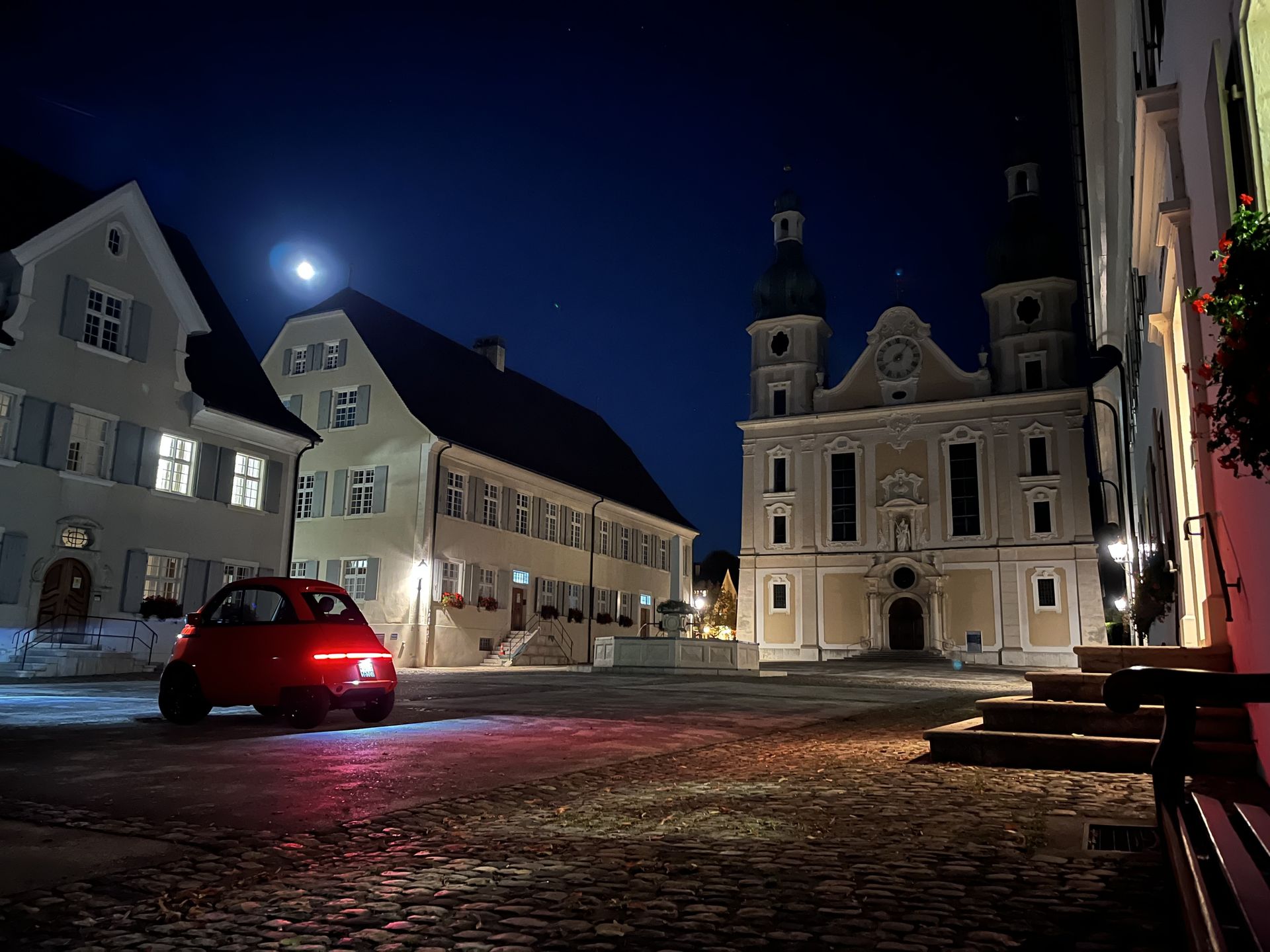 Nächtliche Aufnahme des Domplatzes in Arlesheim mit der beleuchteten Domkirche, historischen Gebäuden und einem roten Auto im Vordergrund.